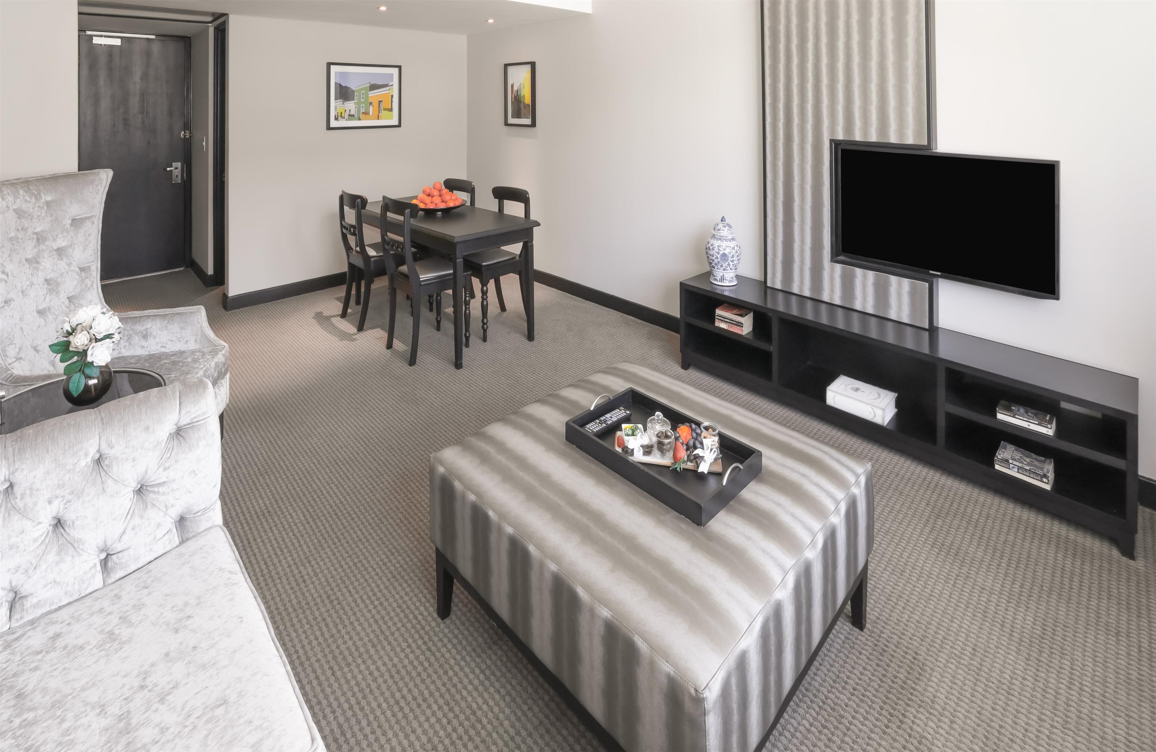 Una sala moderna con un sofá gris, una mesa de comedor con sillas, un televisor de pantalla plana y una bandeja con varios objetos sobre un otoman.