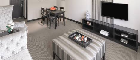 Una sala moderna con un sofá gris, una mesa de comedor con sillas, un televisor de pantalla plana y una bandeja con varios objetos sobre un otoman.