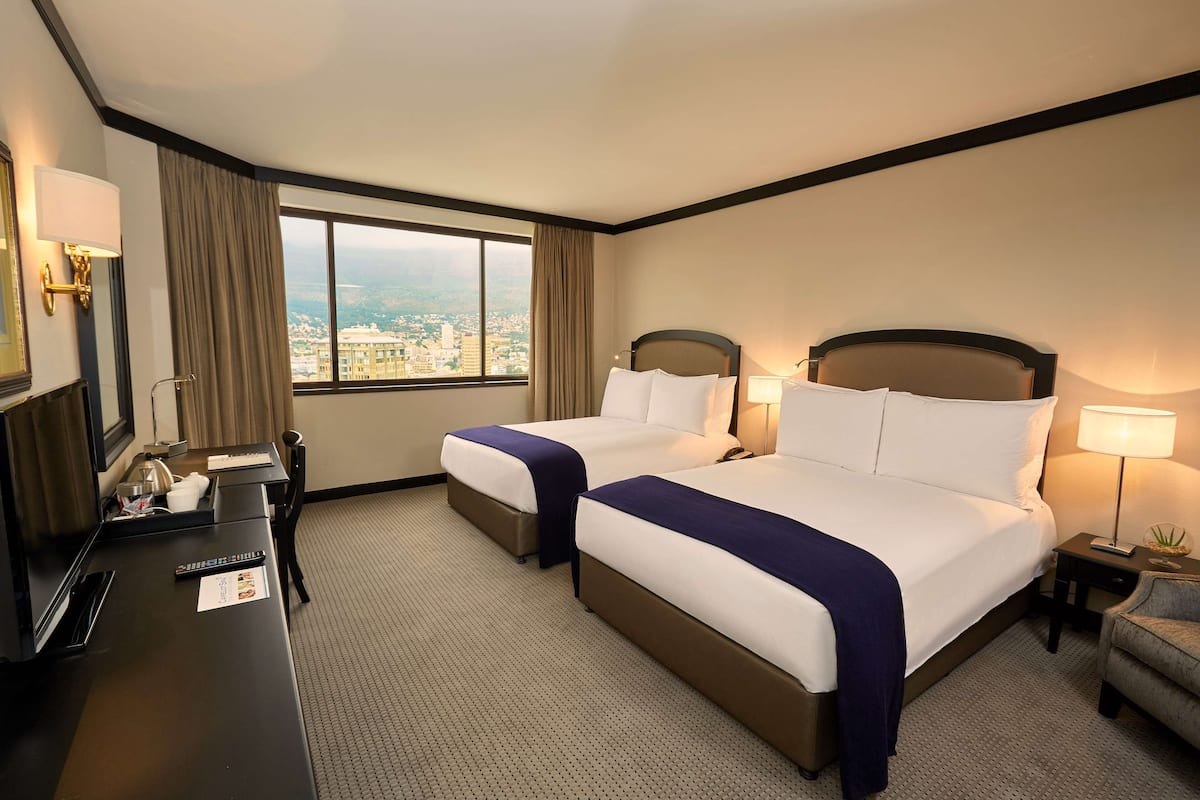 A hotel room with two beds, a desk, a chair, a nightstand, and a window with a city view.