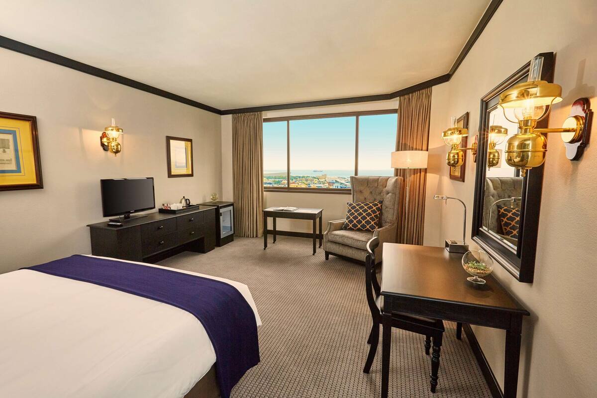 A hotel room with a bed, a desk, a chair, a TV, and a large window offering a city view.