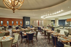 A spacious hotel dining area with multiple tables and chairs, a central chandelier, and a kitchen area in the background.