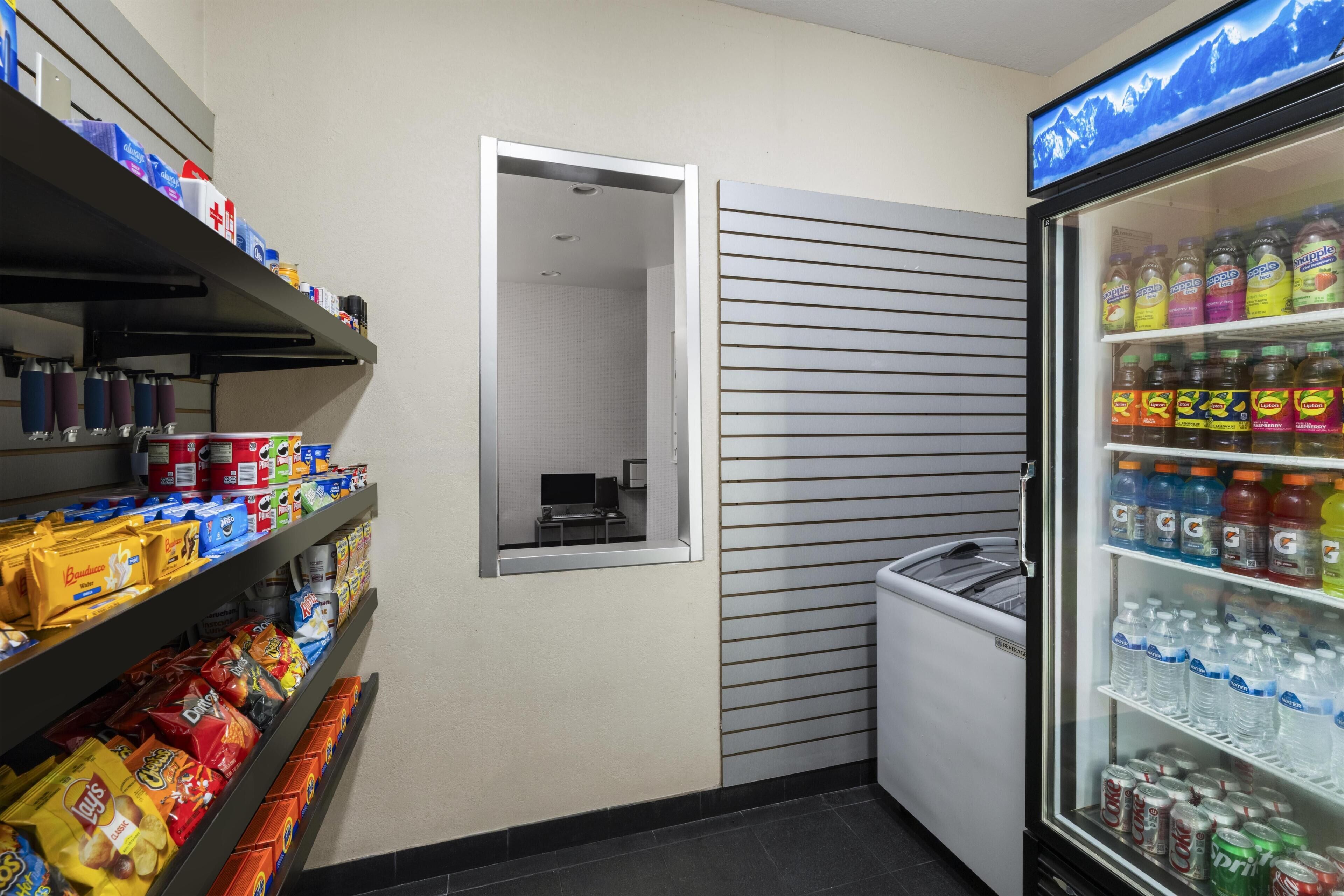 A corner of a hotel room with shelves stocked with snacks, a refrigerator with beverages, and a small window.