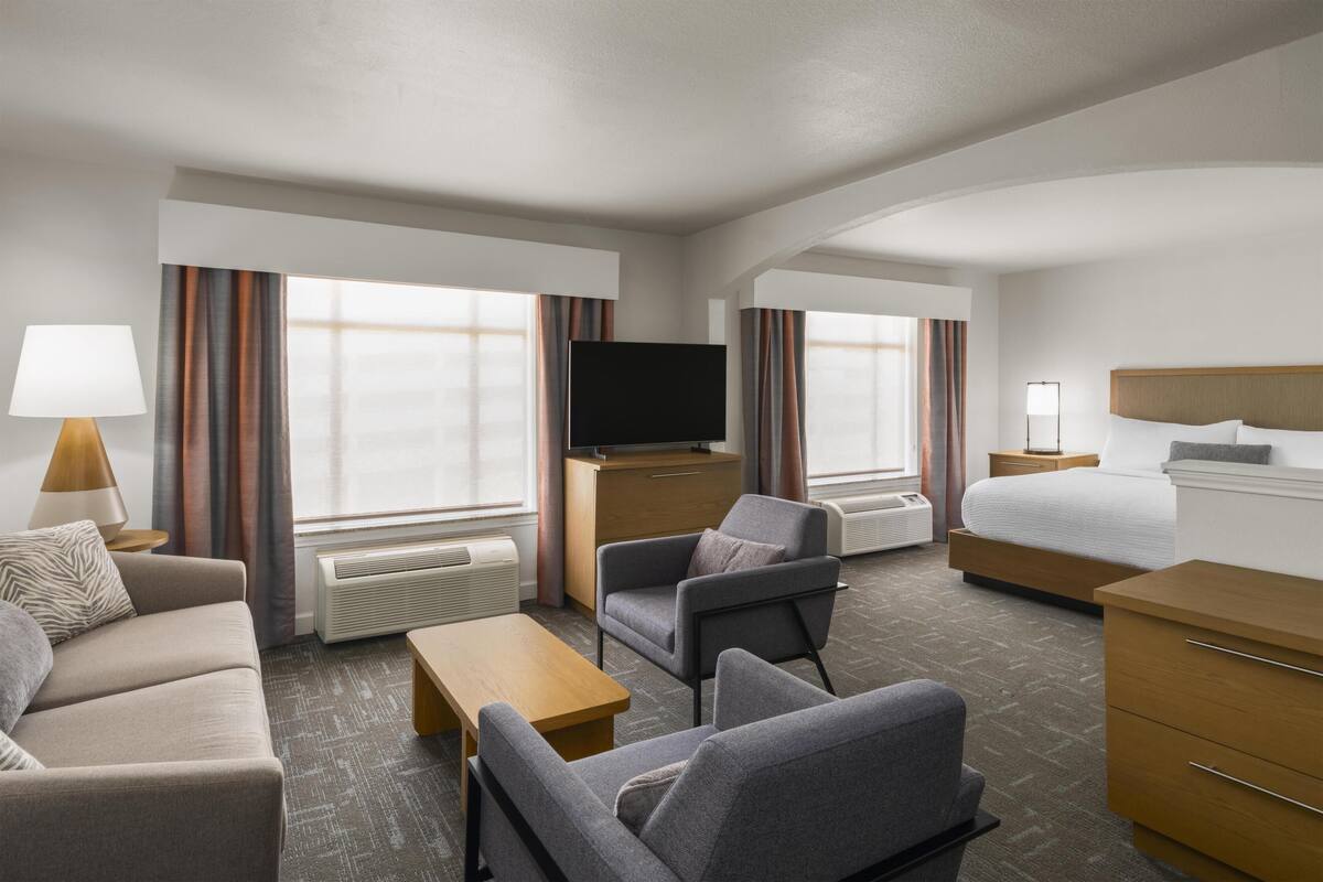 A hotel room with a sofa, armchair, bedside table, TV, bed, and a desk with a lamp.