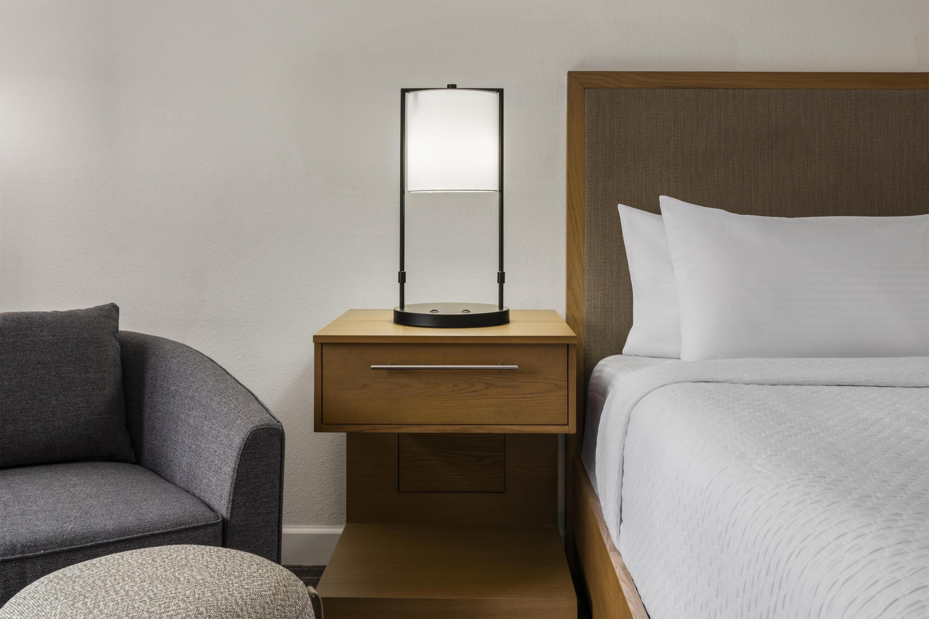 Ein Hotelzimmer mit einem Bett, einem Nachttisch mit Lampe, einem Sessel und einem kleinen Hocker.