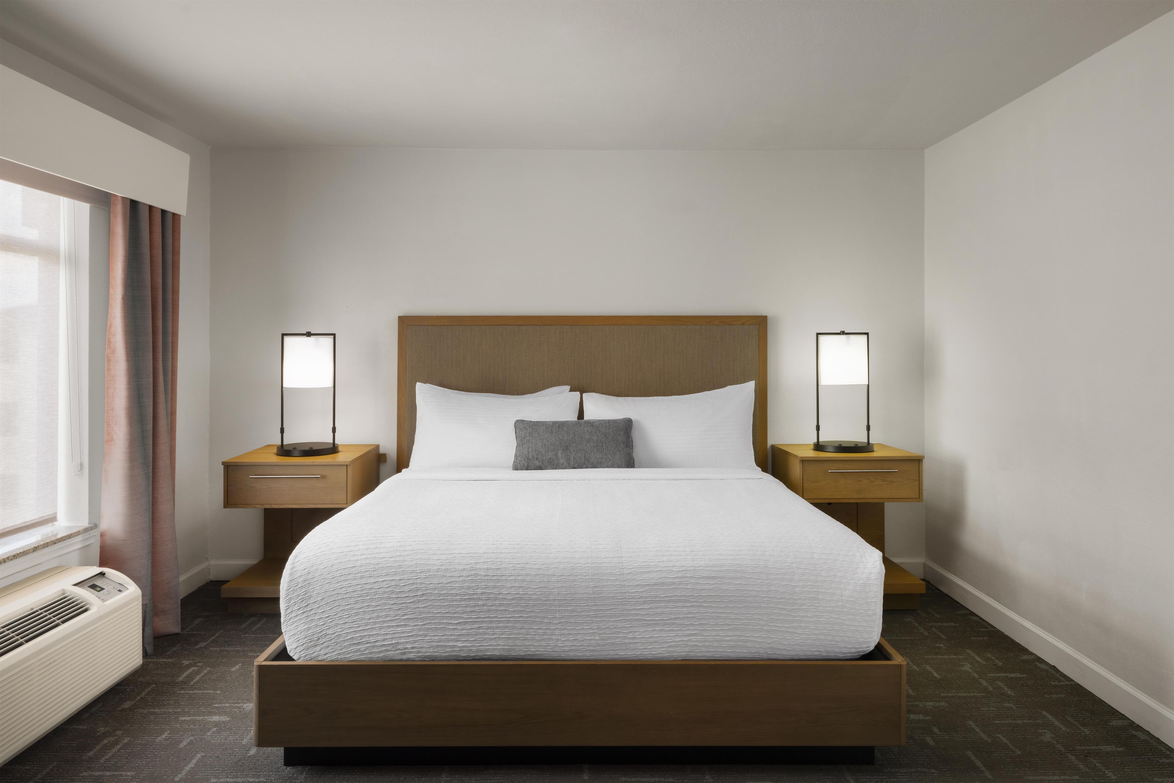 Una habitación de hotel con una cama grande, dos mesitas de noche, un radiador fijado a la pared y una ventana con cortinas.