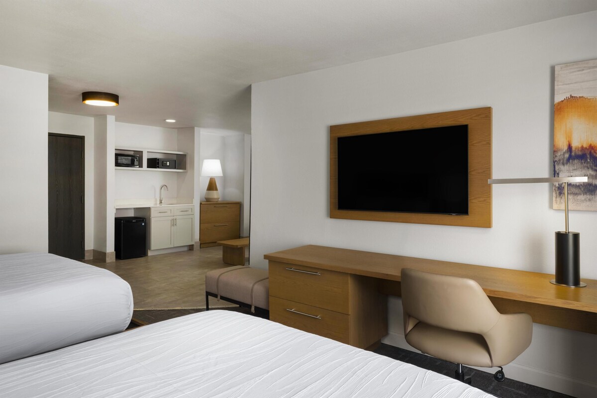 Una camera d'hotel con due letti, una scrivania, una sedia, una televisione, una lampada e un angolo cottura.