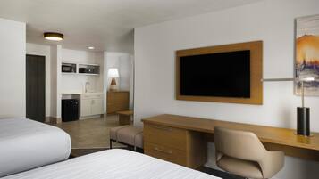 A hotel room with two beds, a desk, a chair, a TV, a lamp, and a kitchen area.