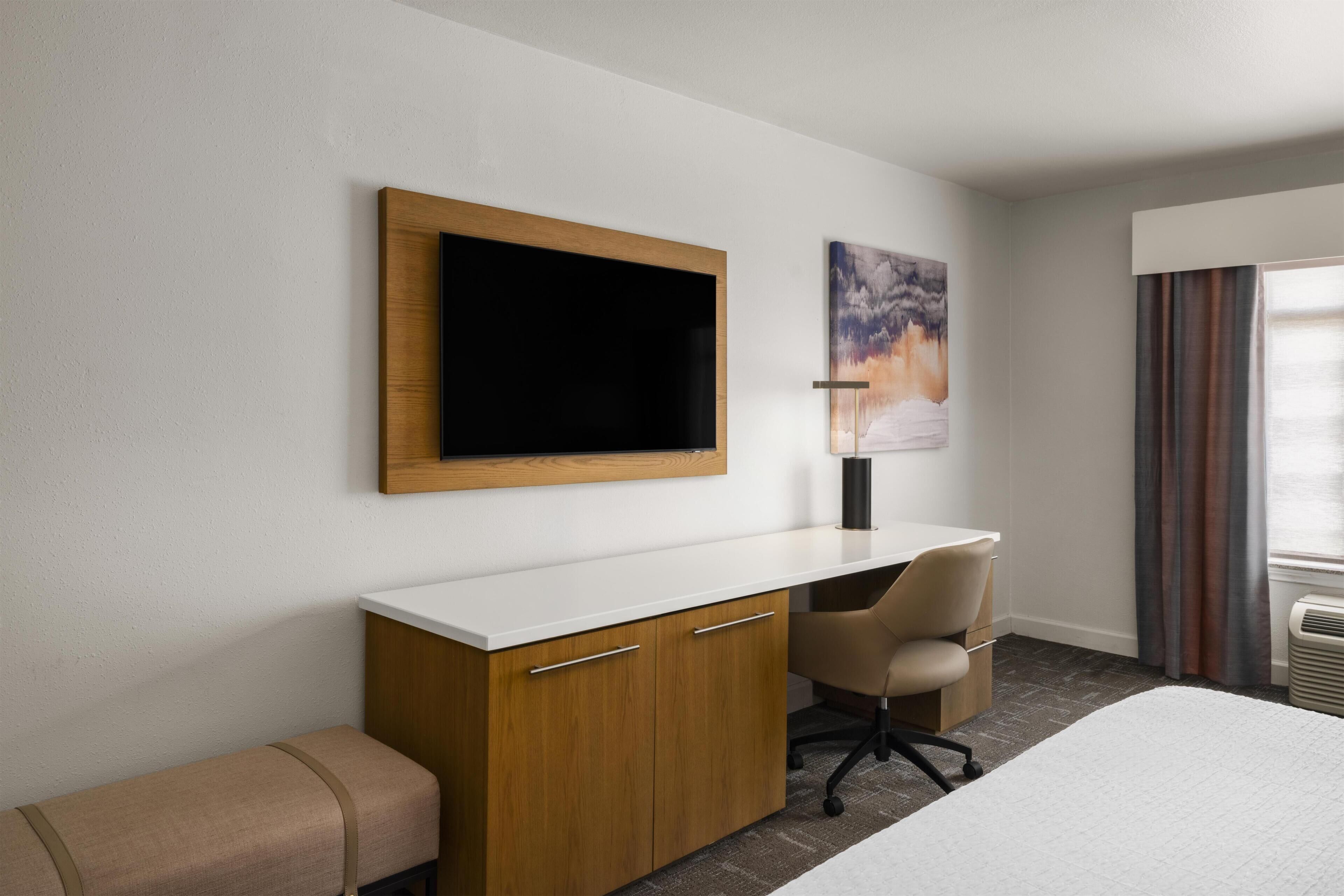 A hotel room with a bed, a desk, a chair, a TV, and a painting on the wall.