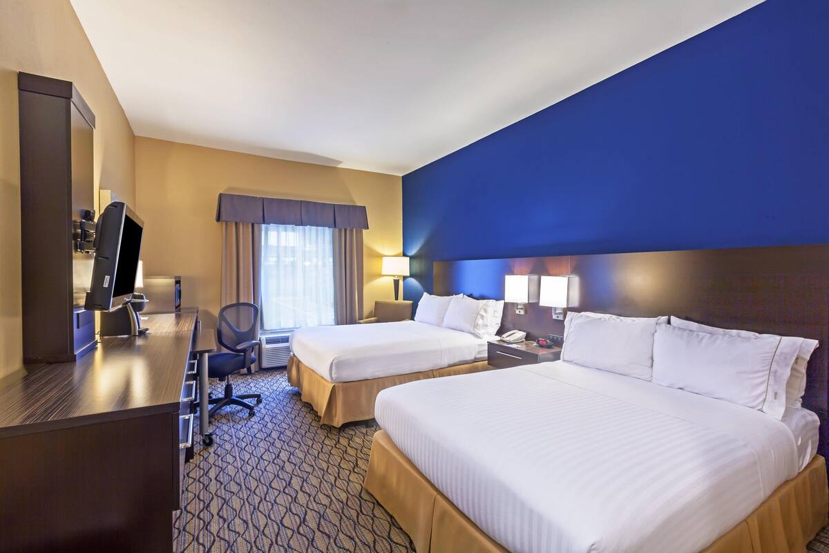 A hotel room with two beds, a desk with a computer, a chair, a lamp, and a large window with curtains.