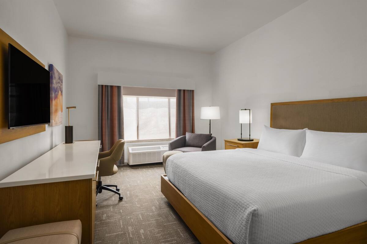 A hotel room with a bed, a desk, a chair, a TV, a lamp, and a window with curtains.
