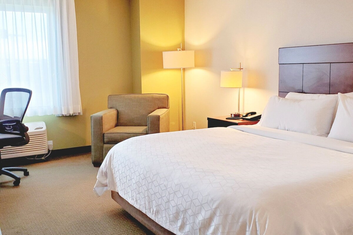 A hotel room with a large bed, a chair, a desk with a laptop, and a lamp.