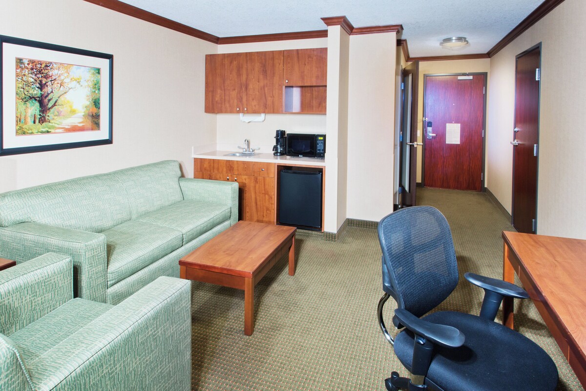 A room with a green sofa, a wooden table, a blue office chair, a kitchenette with a microwave, and a framed picture on the wall.