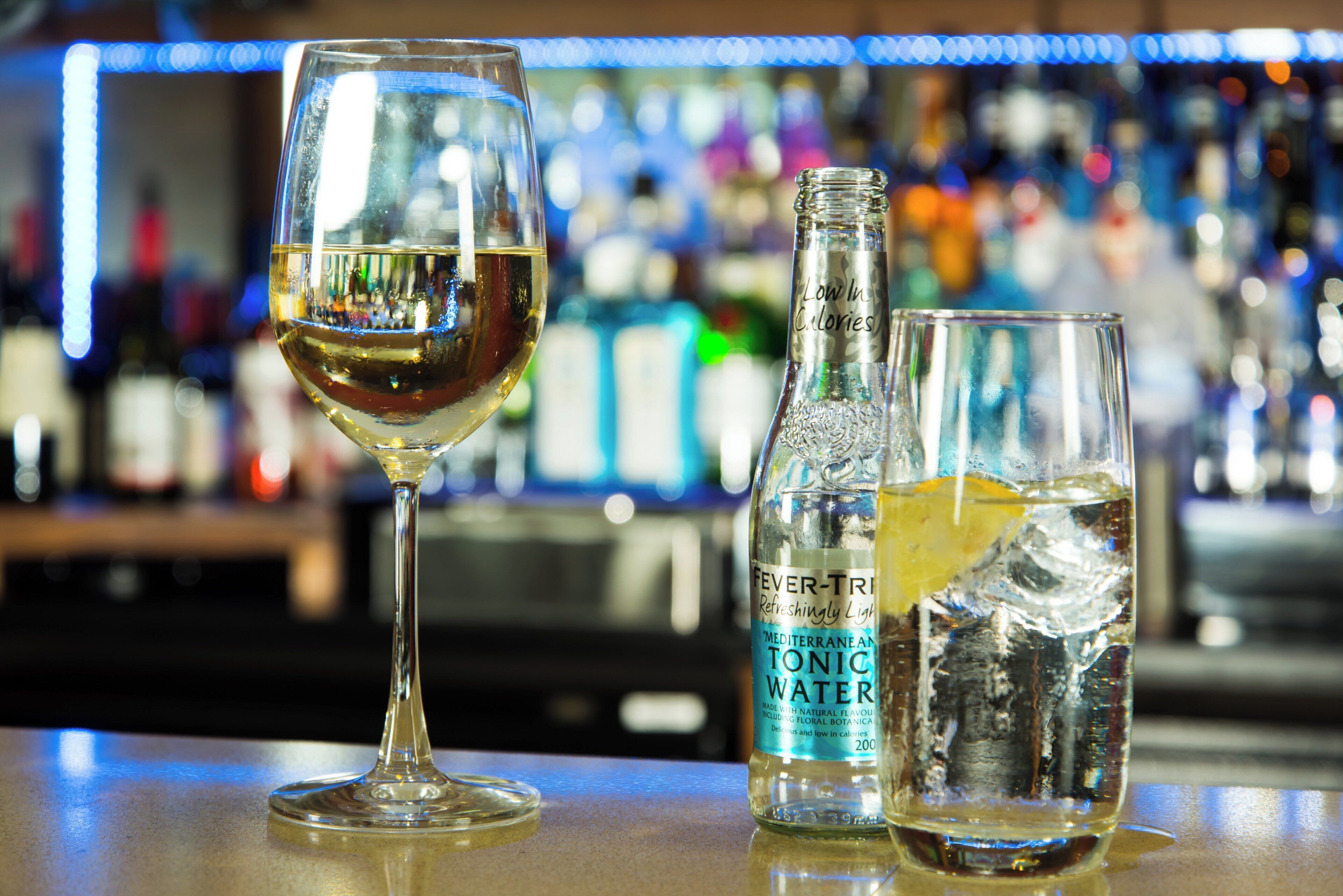 Un bar con una copa de vino blanco, una botella de agua tónica y un vaso con una rodaja de limón.