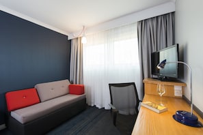 A hotel room with a sofa, a chair, a desk with a television, and a window with curtains.