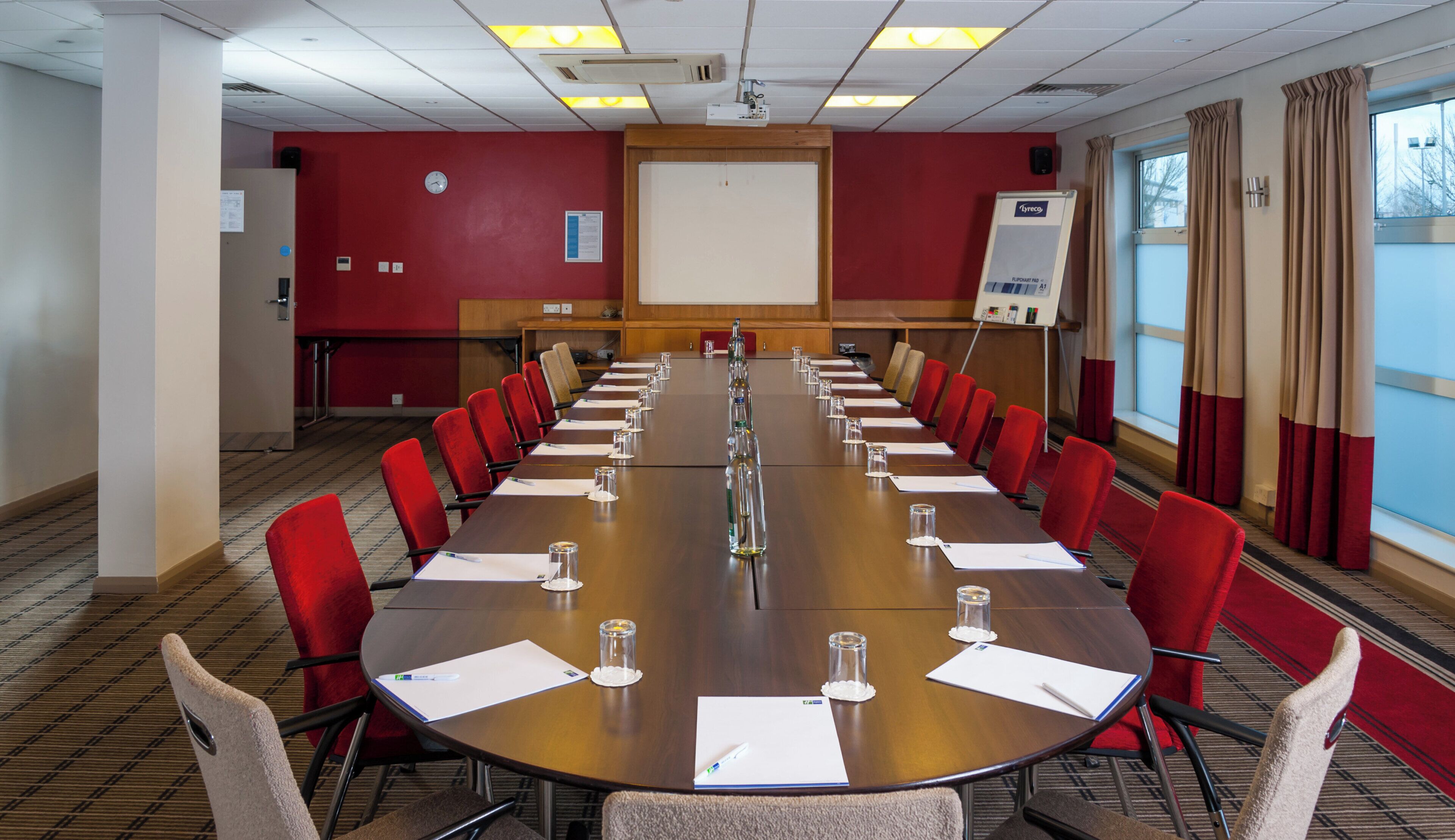 A conference room with a long table, red chairs, and a projector screen.