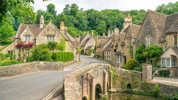 A stone bridge over a river, surrounded by traditional stone houses with chimneys, a curved road, and lush greenery.