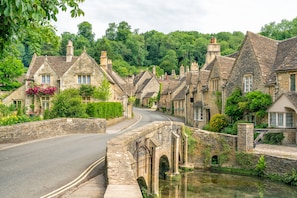 A stone bridge over a river, surrounded by traditional stone houses with chimneys, a curved road, and lush greenery.