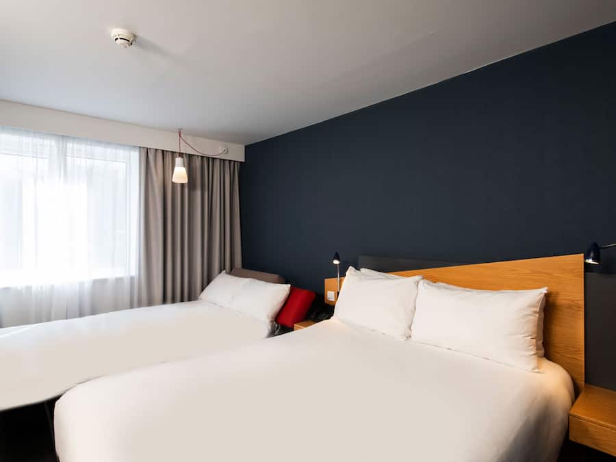 Habitación de hotel con dos camas, cabecero de madera, mesita de noche con lámpara y ventana con cortinas.