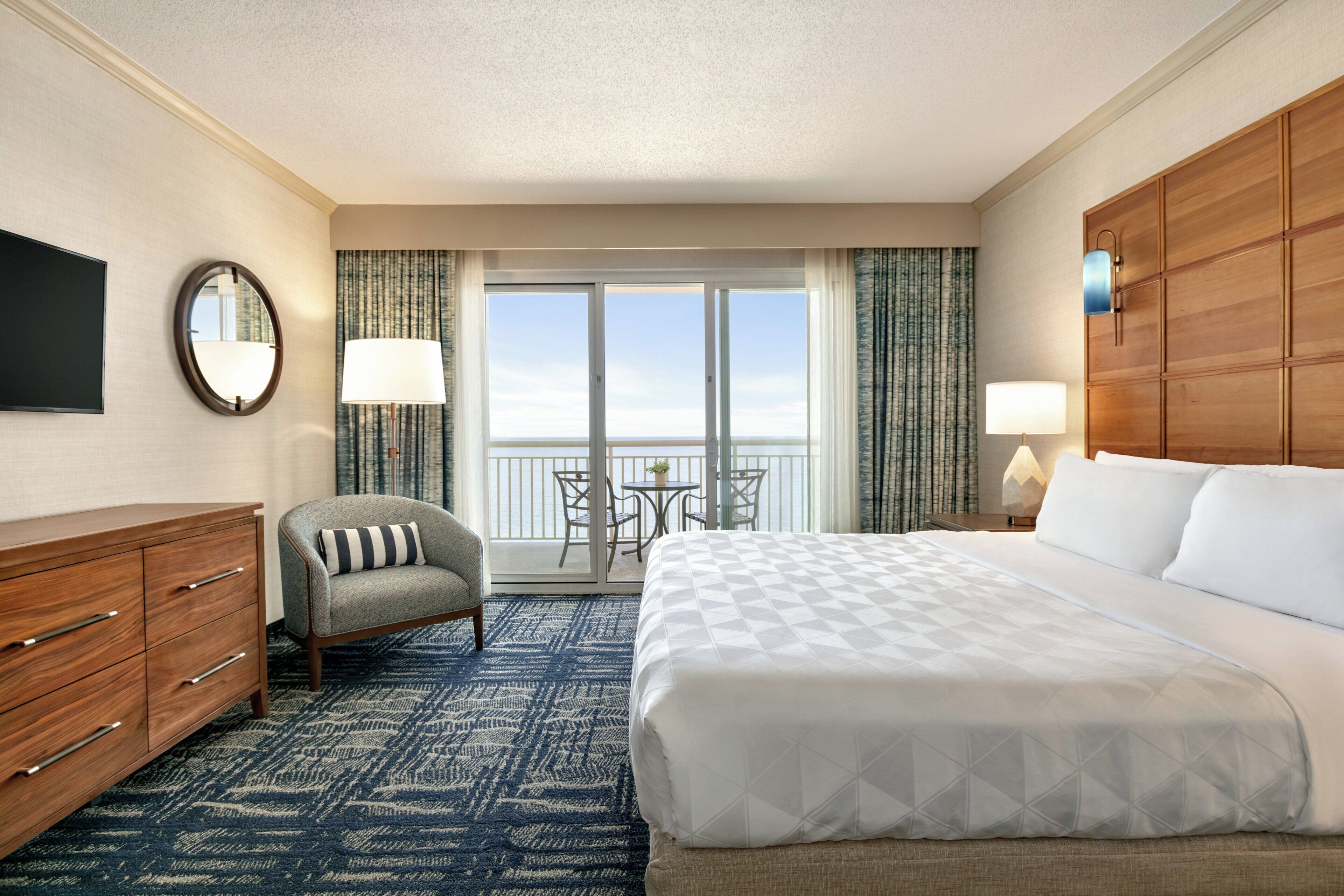 A hotel room with a large bed, a desk, a chair, a TV, and a balcony with a view.
