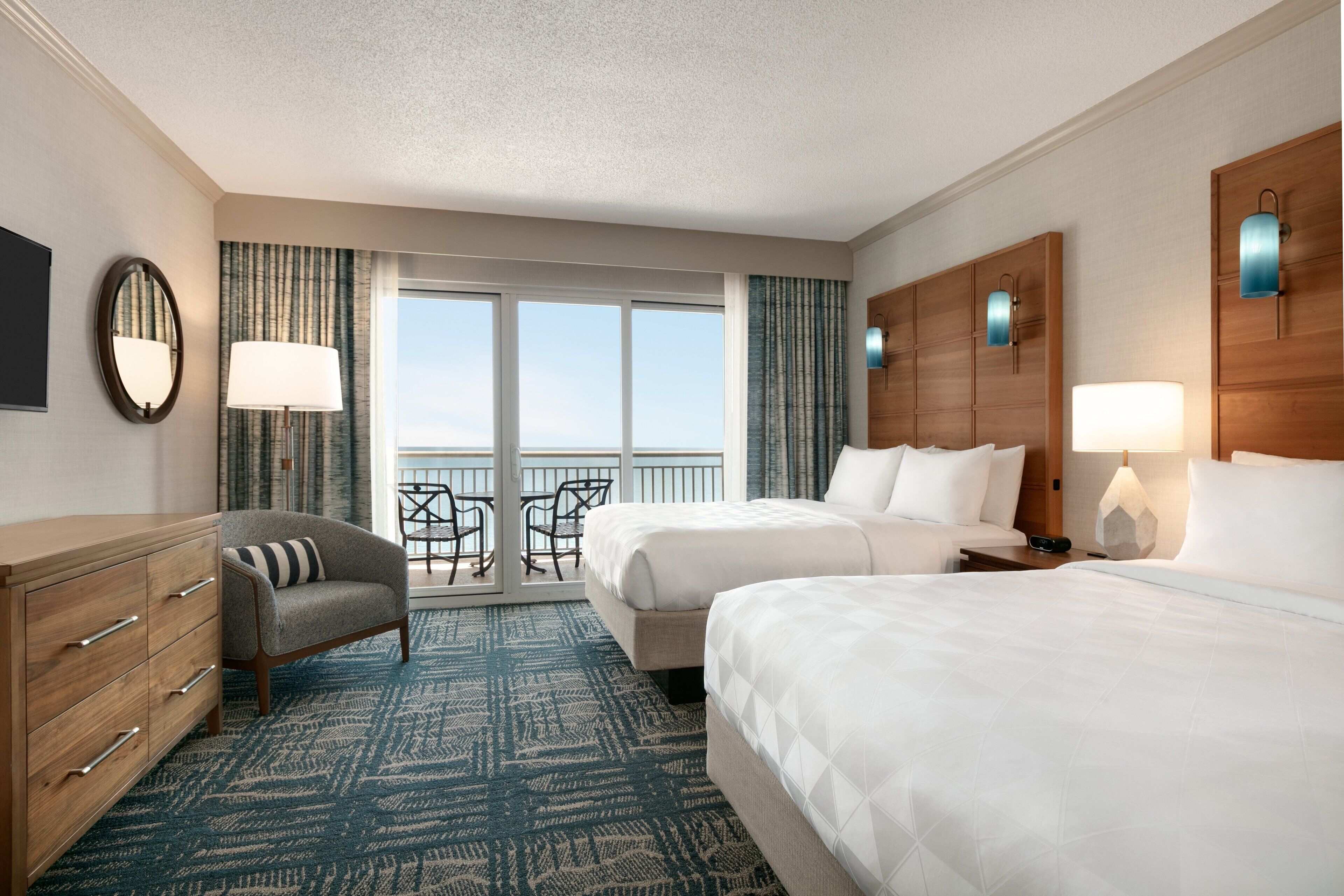 A hotel room with two beds, a wooden dresser, a chair, a balcony with a view, and a large window.