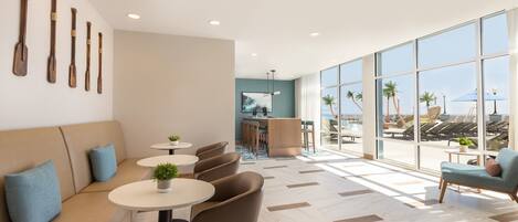 Ein moderner Lounge-Bereich mit einer Couch, runden Tischen, Sesseln, einer Bar und großen Fenstern mit Blick auf Palmen und einen Strand.
