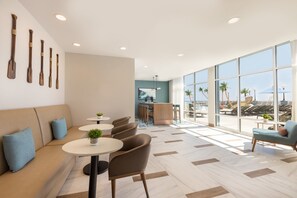 A modern lounge area with a sofa, round tables, and chairs, a bar area, and large windows with a view of palm trees and a beach. - Holiday Inn & Suites Ocean City by IHG (Ocean City)