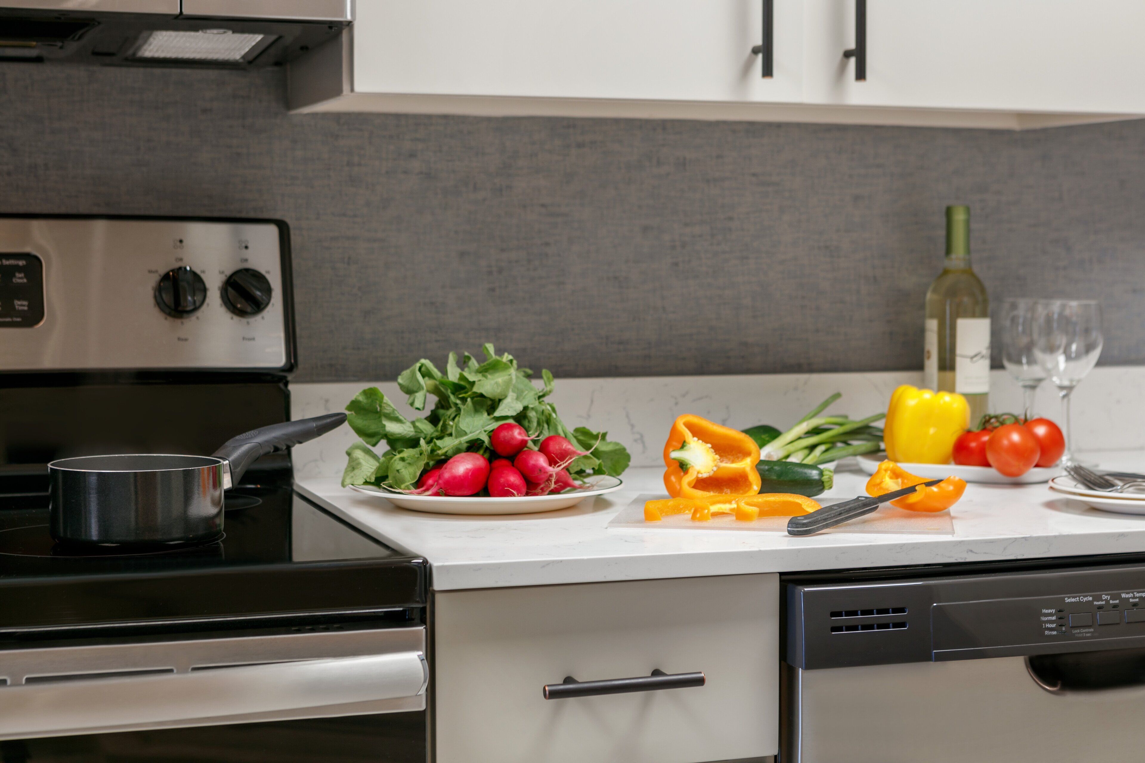 Une cuisine équipée d’une cuisinière, d’un lave-vaisselle et d’un plan de travail sur lequel on trouve des légumes, un couteau et une bouteille de vin.