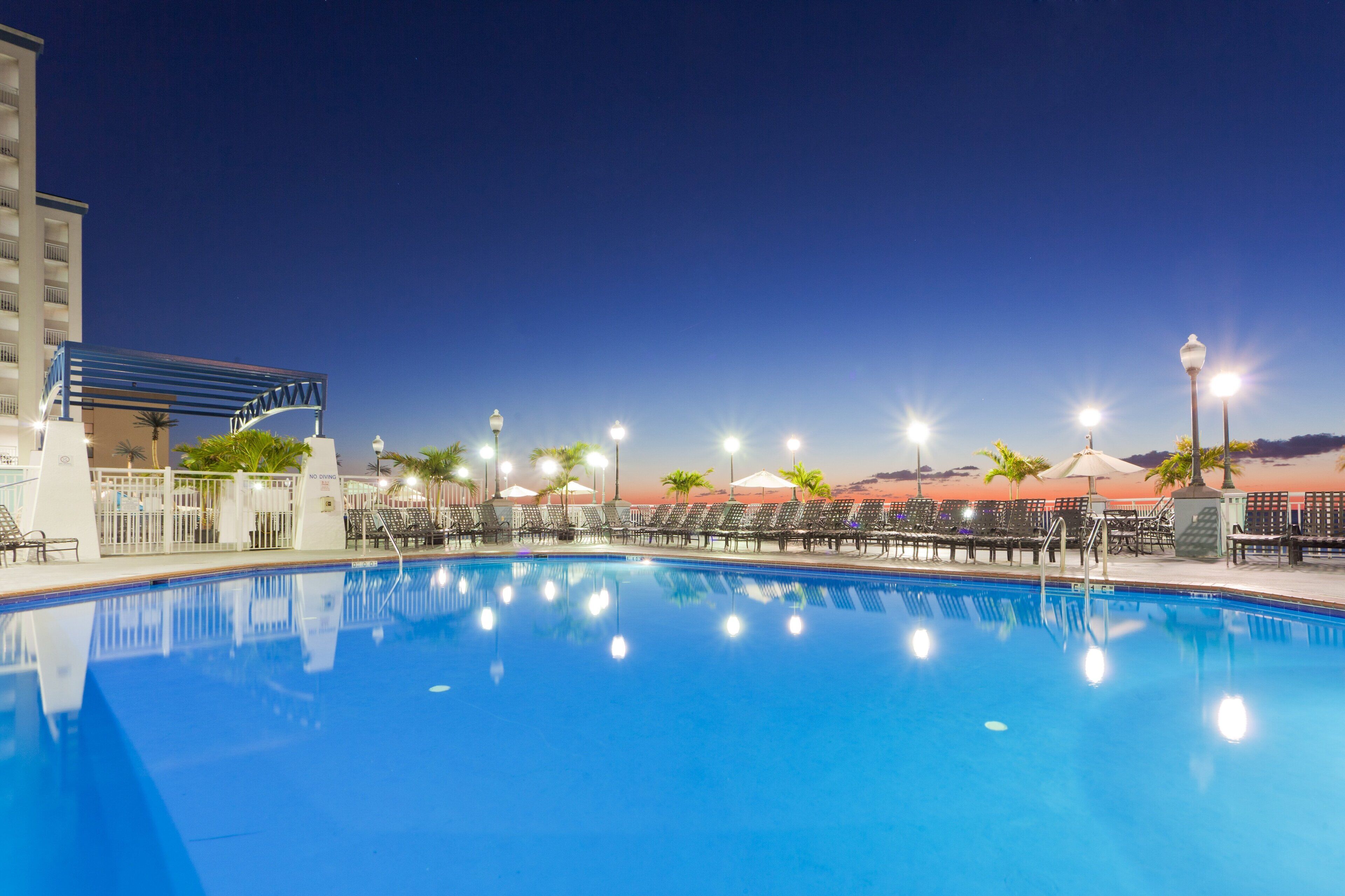 A swimming pool with lounge chairs and umbrellas, surrounded by a fence and palm trees.