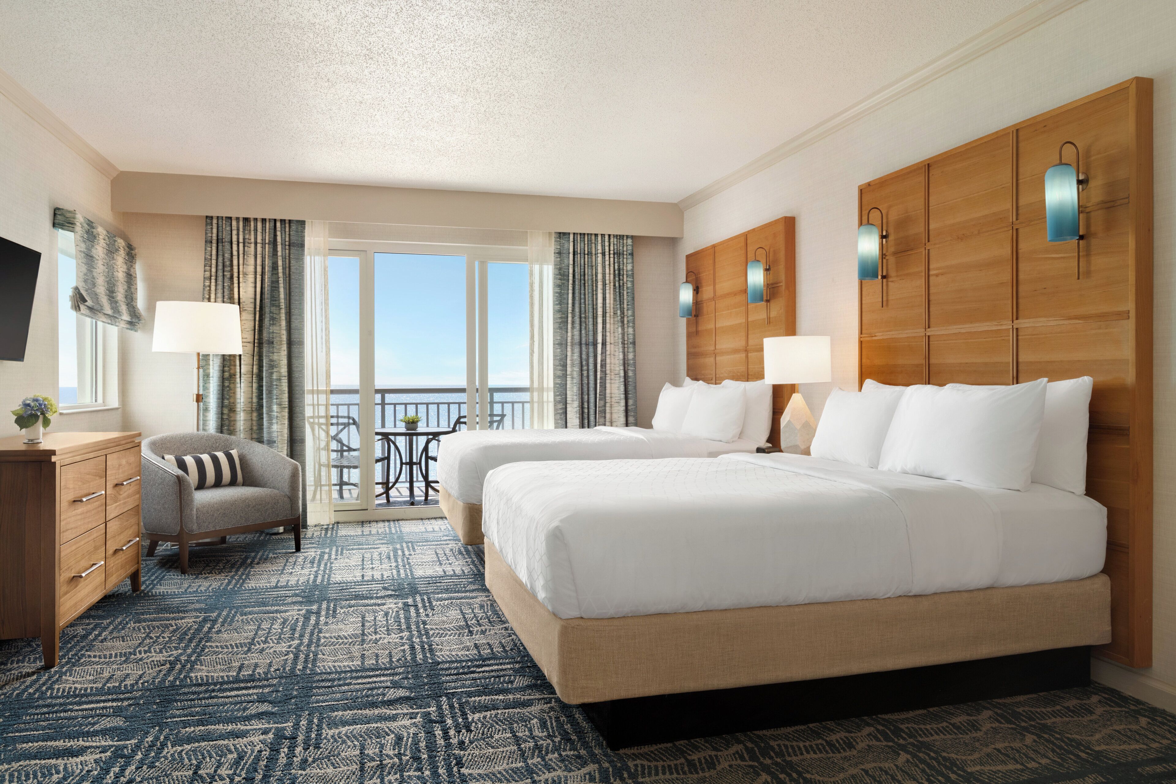 A hotel room with two beds, a desk, a chair, and a balcony with a view.