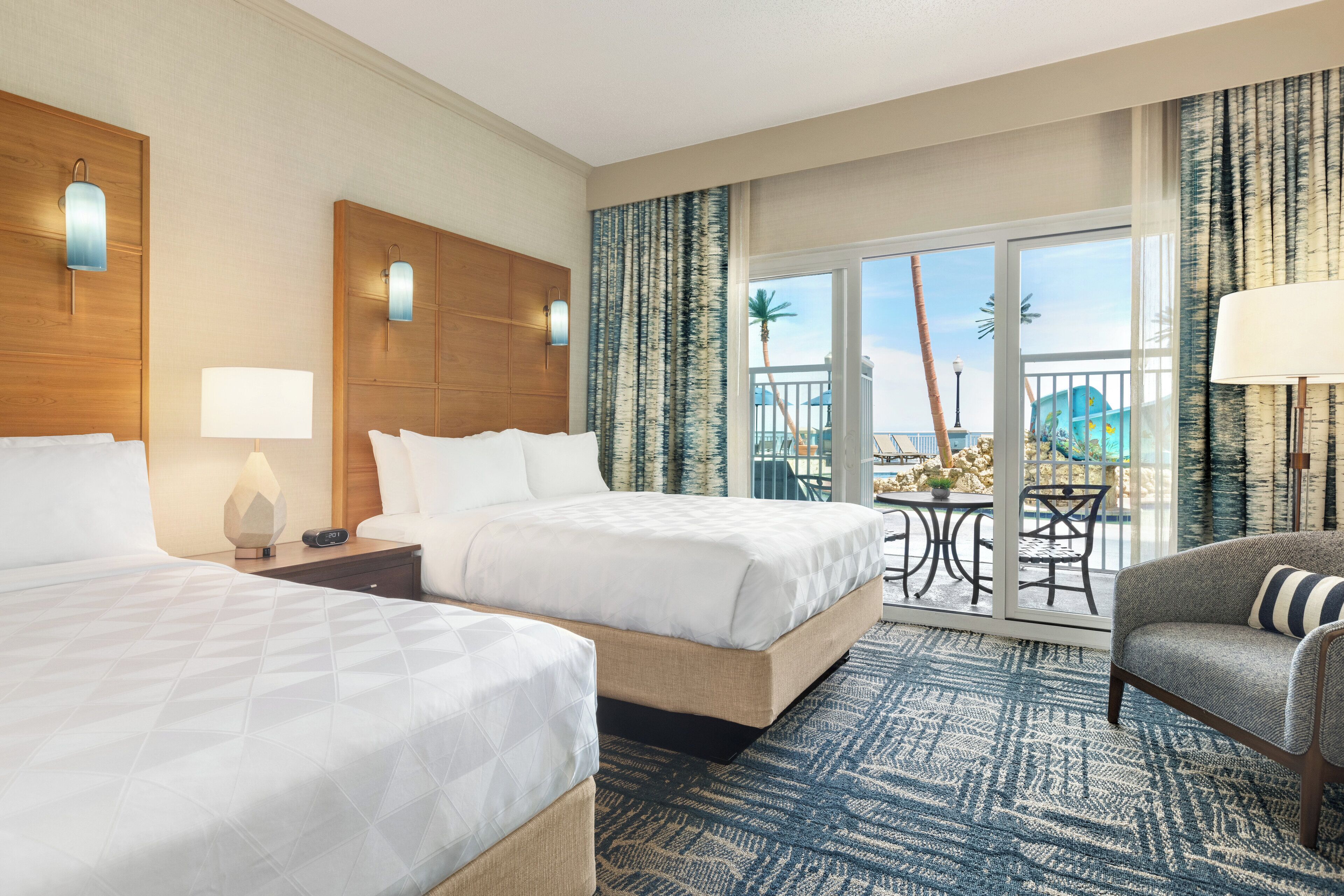 Habitación de hotel con dos camas, un sofá, un balcón y vistas a palmeras, además de una alfombra estampada.