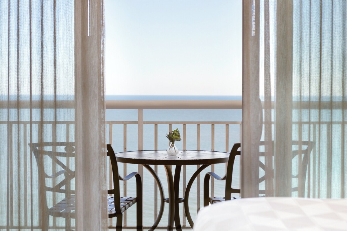 A balcony with a table and chairs, a small vase with a plant, and a view of the sea.