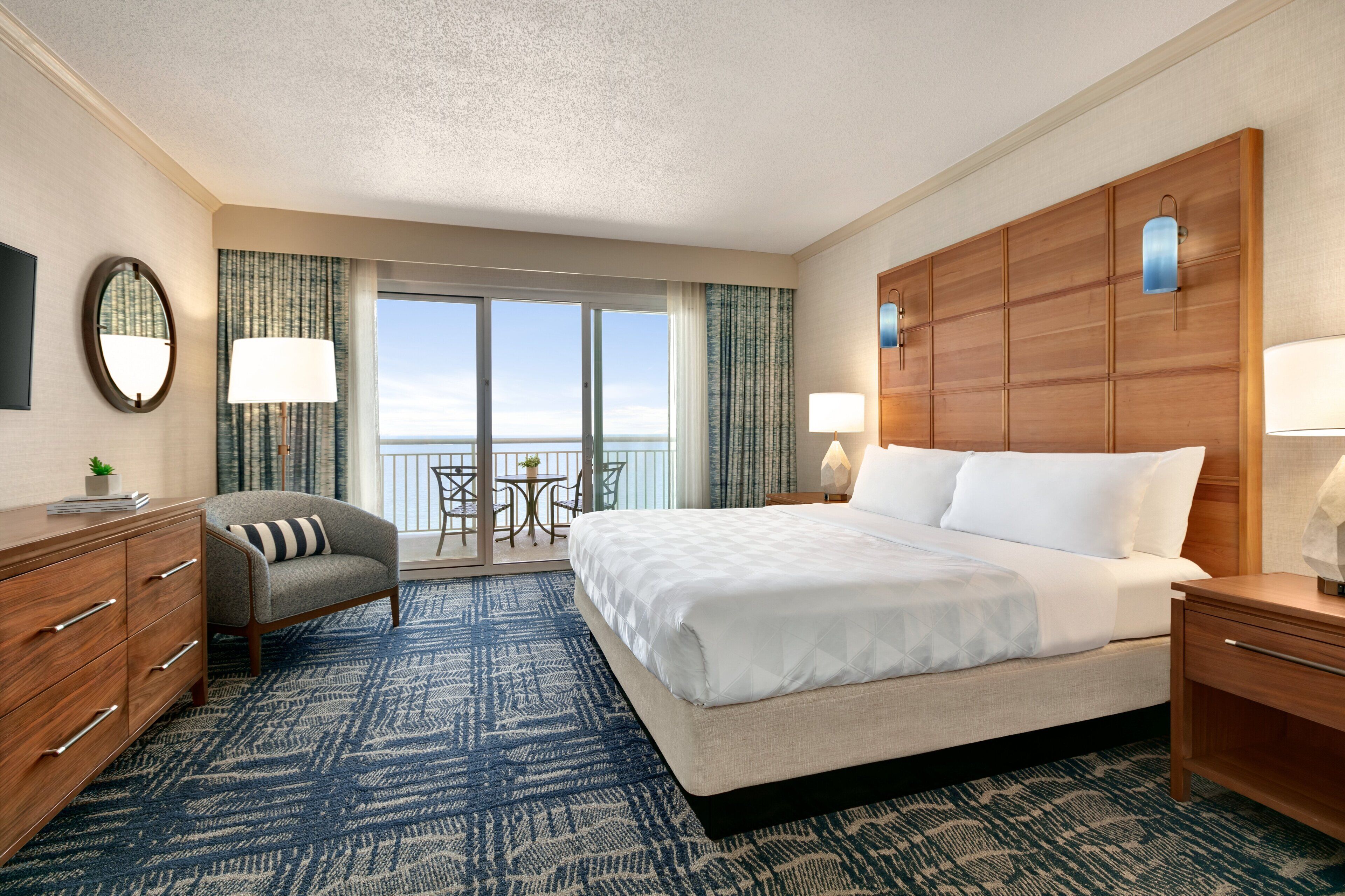 A hotel room with a large bed, a desk, a chair, a small table, and a balcony with a view of the ocean.