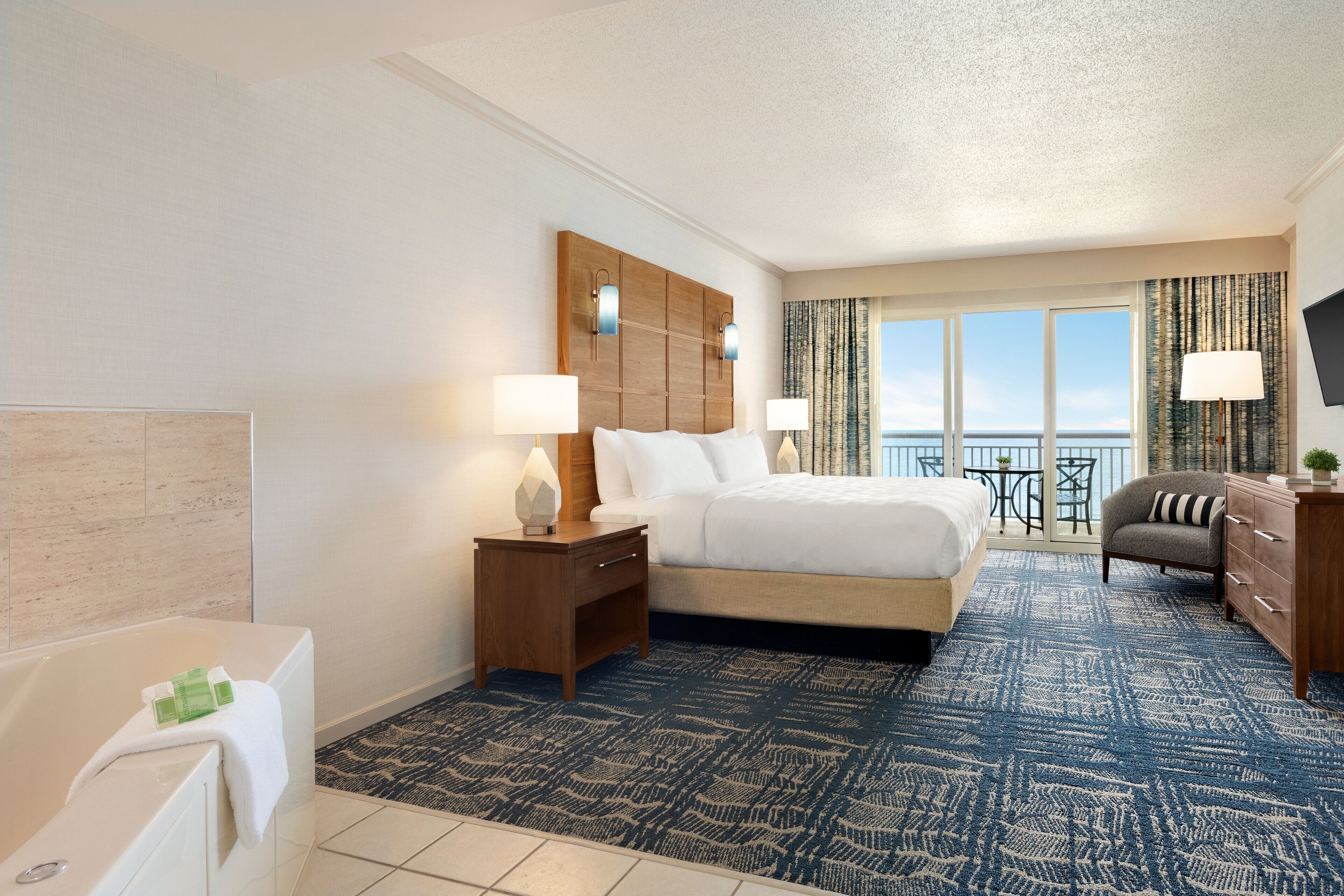 Habitación de hotel con una cama grande, una zona de estar con una silla, una pequeña mesa, un sofá, un televisor y vista al océano.