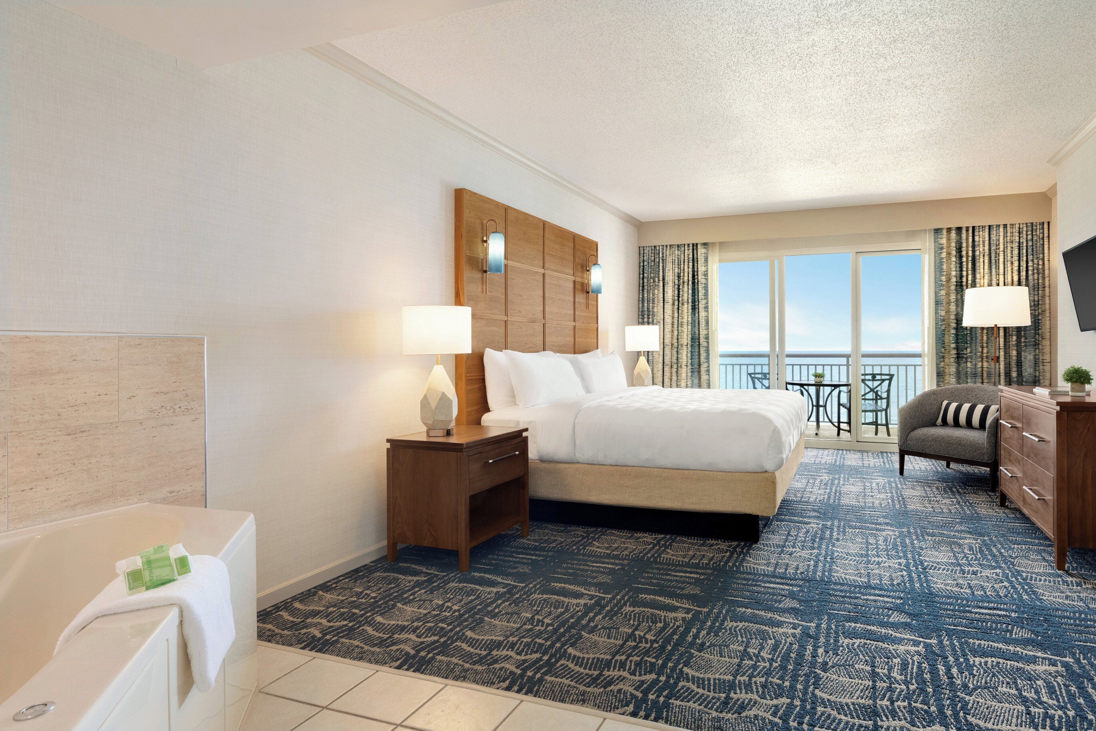 Habitación de hotel con cama, mesita de noche, escritorio, silla, baño y balcón con vistas.