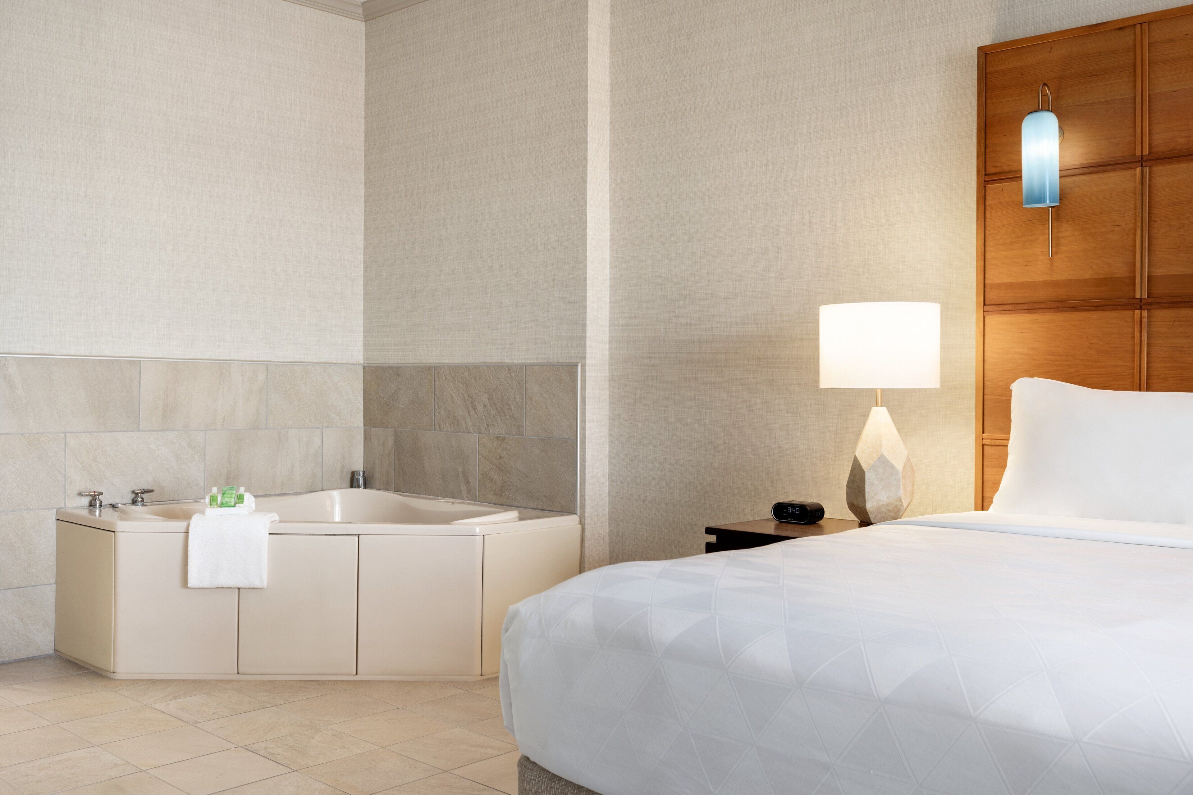 A hotel room with a bed, a nightstand with a lamp, a bathroom with a bathtub, and a wall-mounted light fixture.