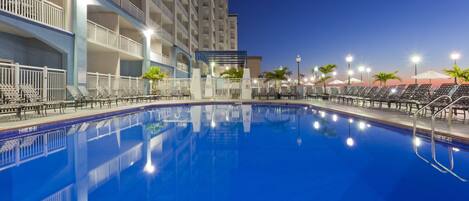 A swimming pool with lounge chairs and umbrellas, surrounded by a multi-story building with balconies.