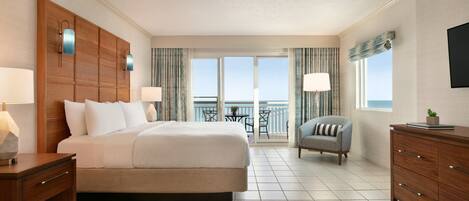 A hotel room with a large bed, a desk with a lamp, a chair, a dresser, and a balcony with a view.
