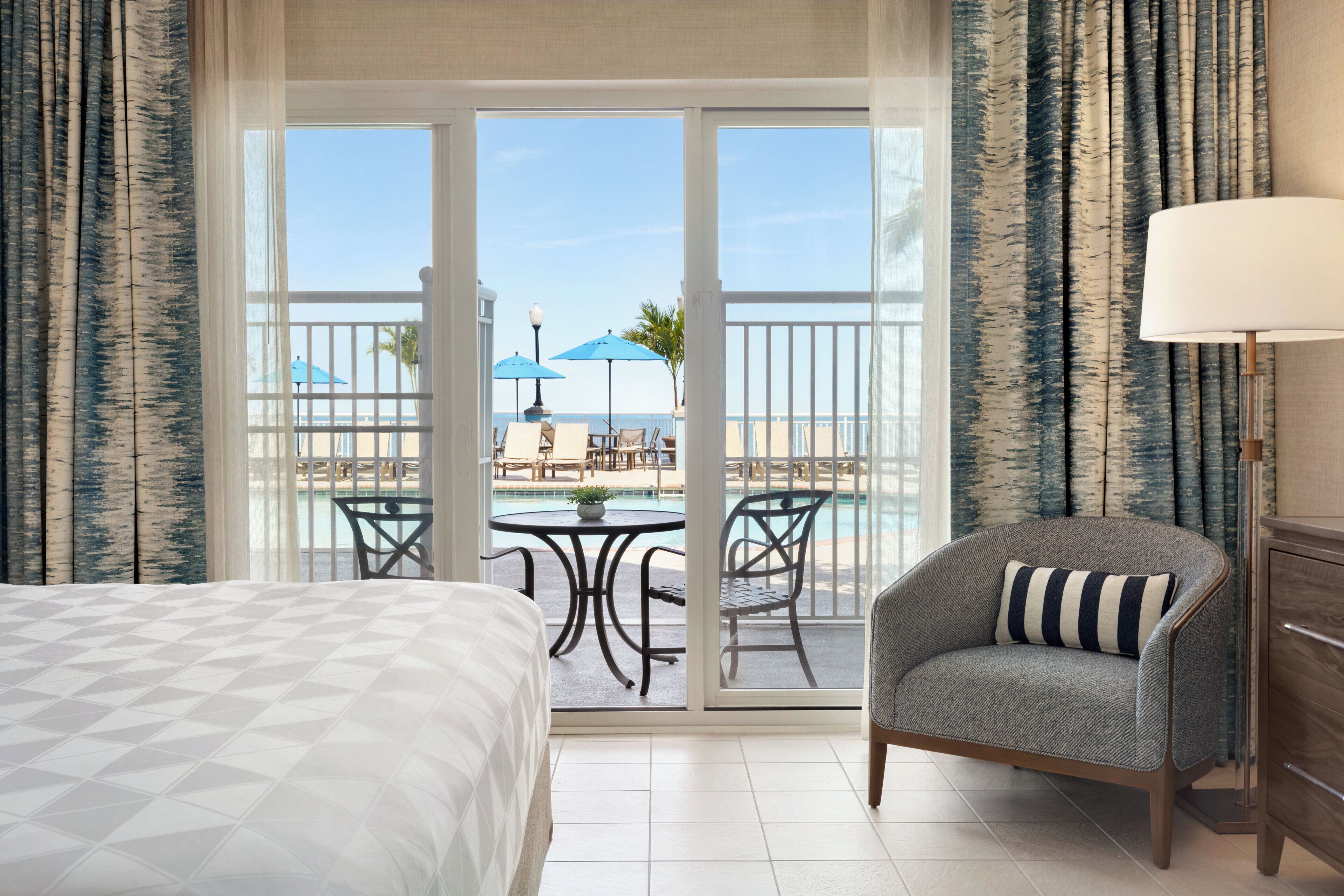 A hotel room with a bed, a chair, a table, and a view of a pool and beach.