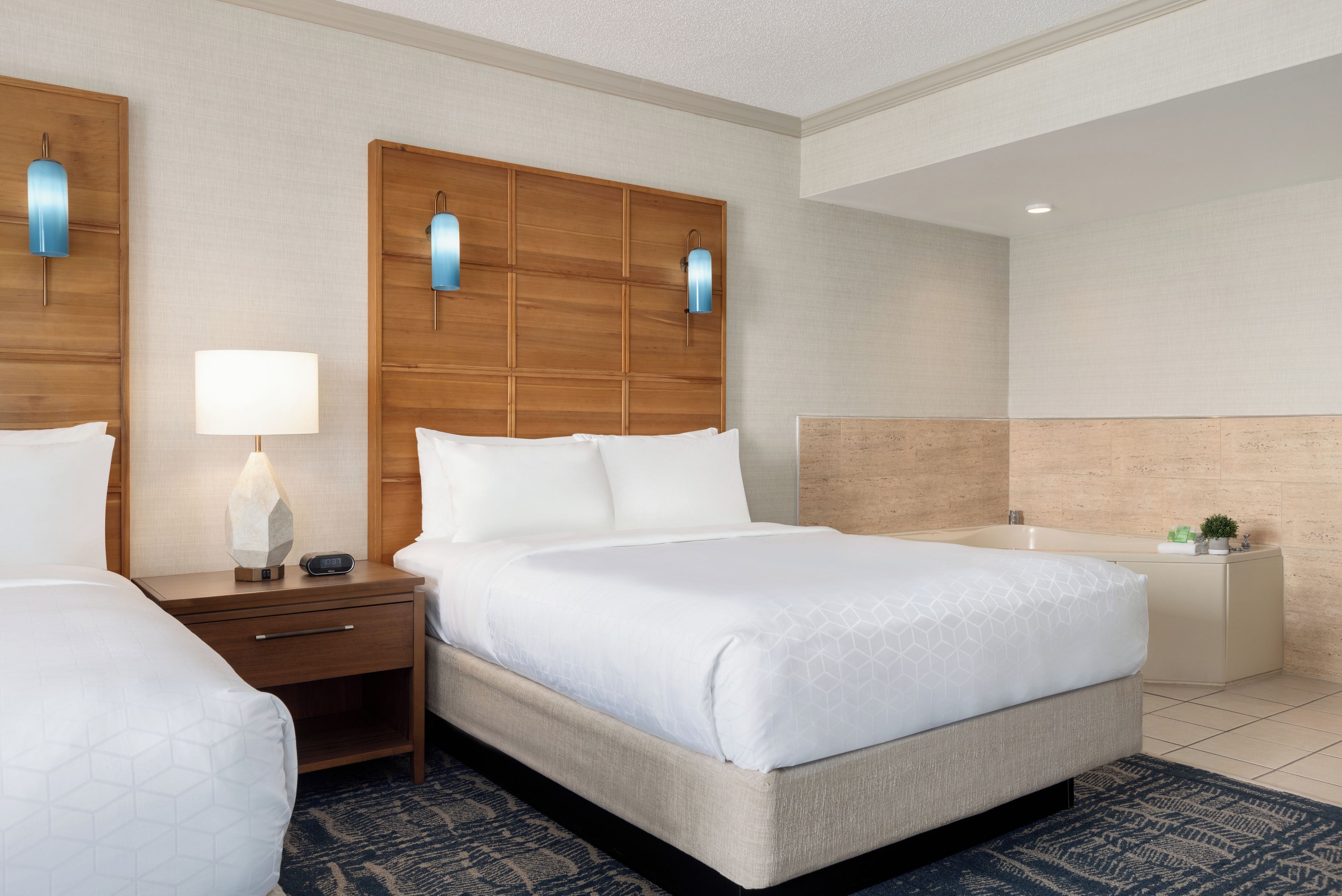 A hotel room with a bed, a nightstand with a lamp, a digital clock, a bedside table, and a bathroom with a bathtub.
