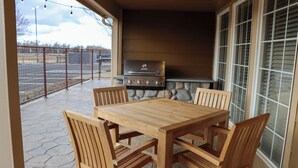 An outdoor patio with a stone floor, a wooden table, and four chairs. A grill is visible in the background.