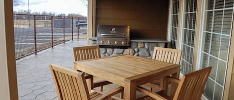 An outdoor patio with a stone floor, a wooden table, and four chairs. A grill is visible in the background.