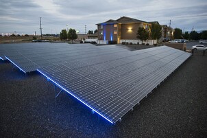 Un extenso campo de paneles solares instalado sobre una superficie plana, probablemente un estacionamiento, con un edificio al fondo.
