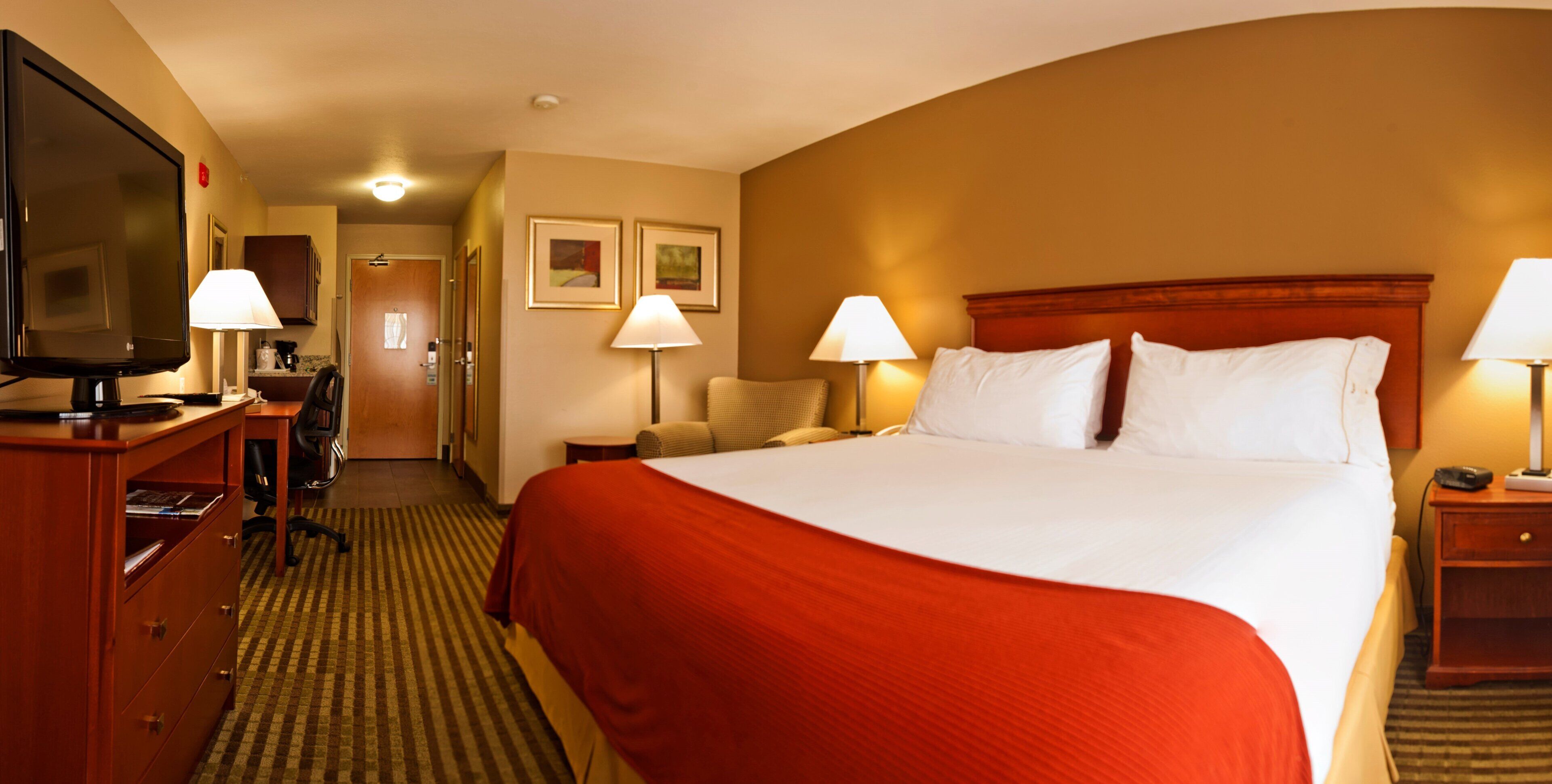 Habitación de hotel con una cama grande, televisor, escritorio con lámpara y puerta que da a un pasillo.
