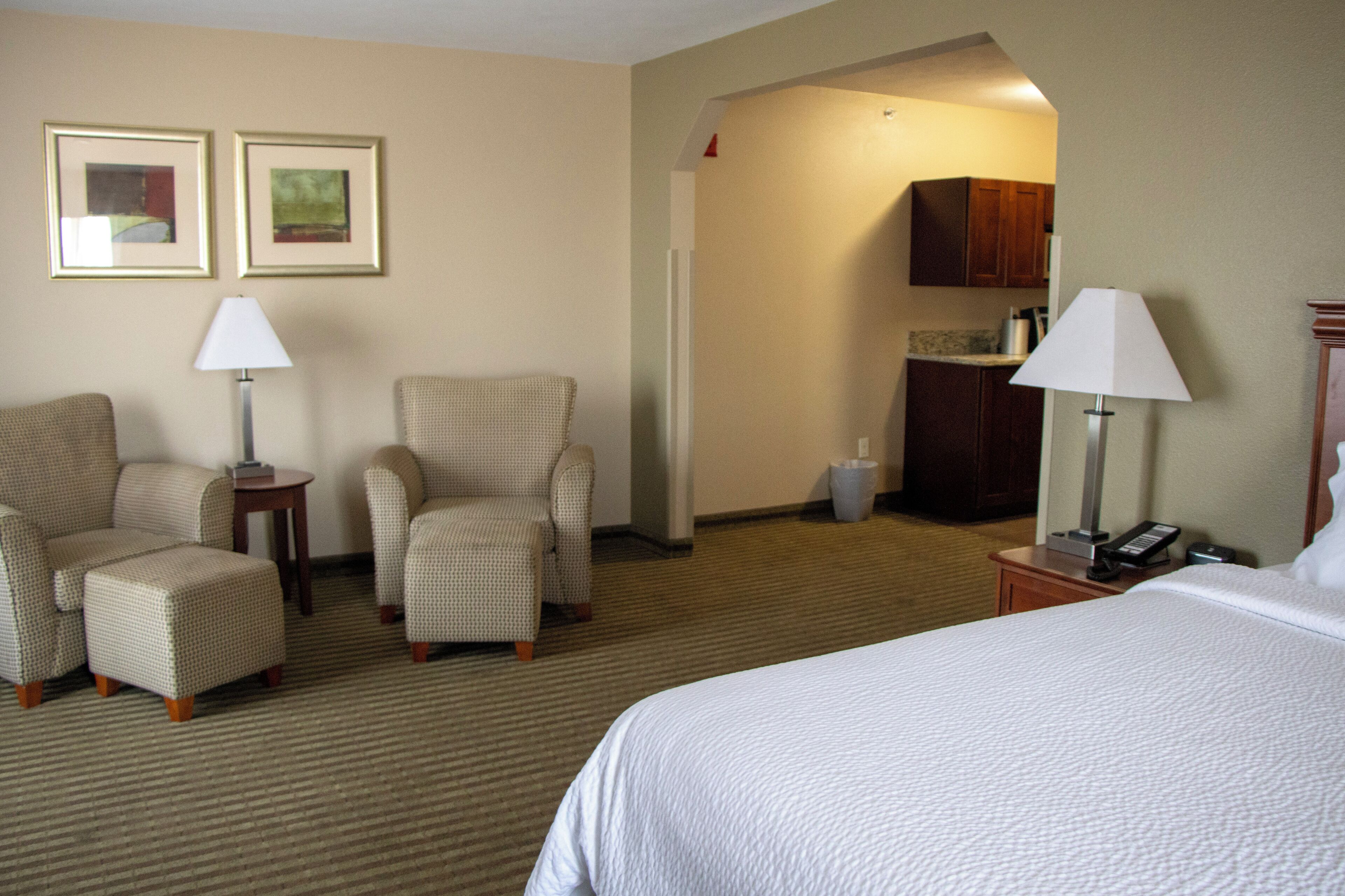 A hotel room with a bed, two armchairs, a small table, a lamp, a picture frame, and a kitchenette area.