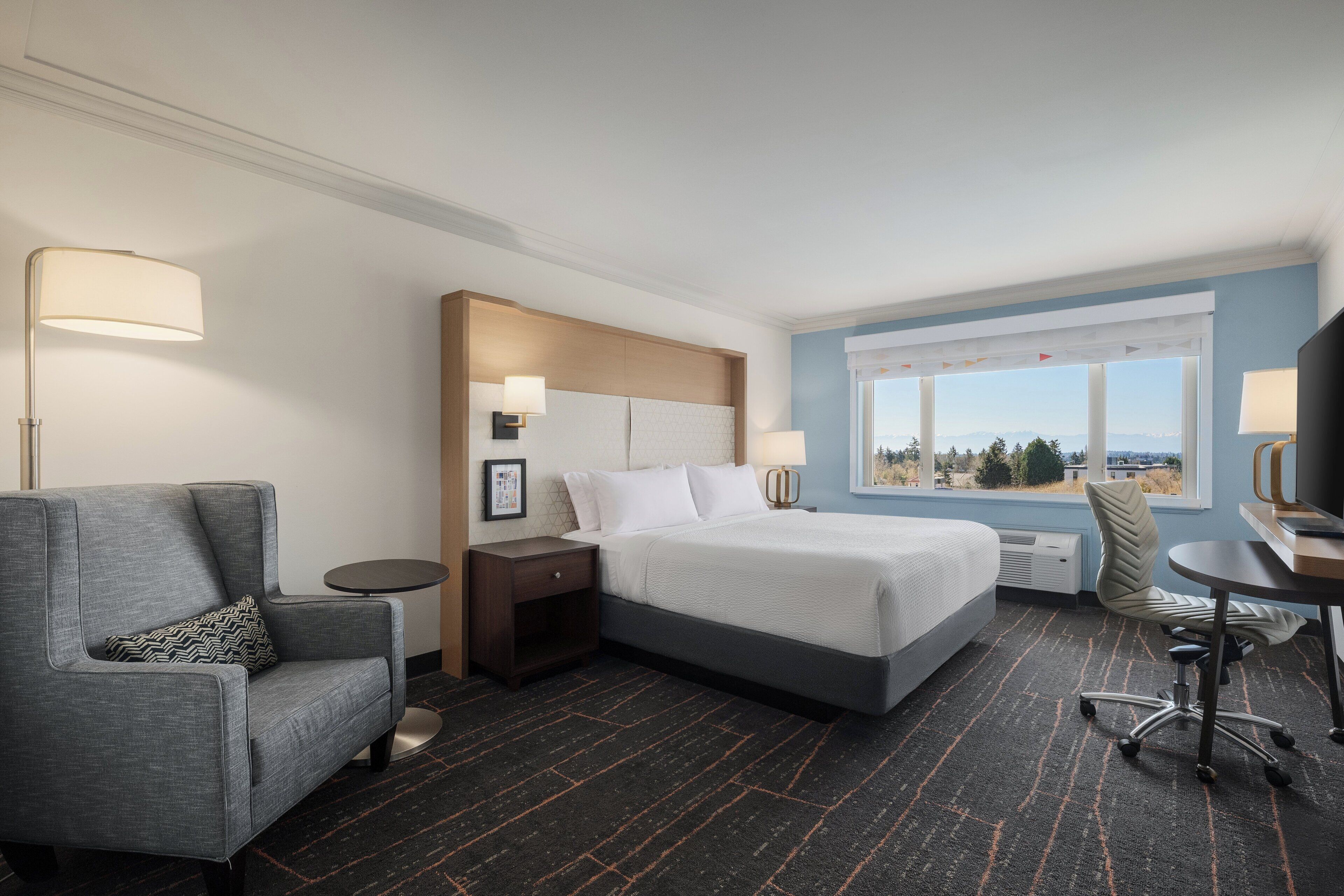 A hotel room with a bed, a gray armchair, a desk, and a view of the outdoors.