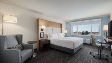 A hotel room with a bed, a gray armchair, a desk, and a view of the outdoors.