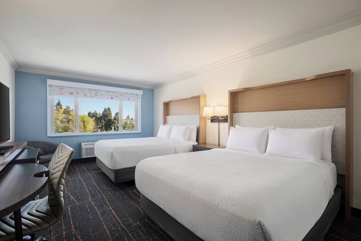 A hotel room with two beds, a desk, and a window with a view of trees.