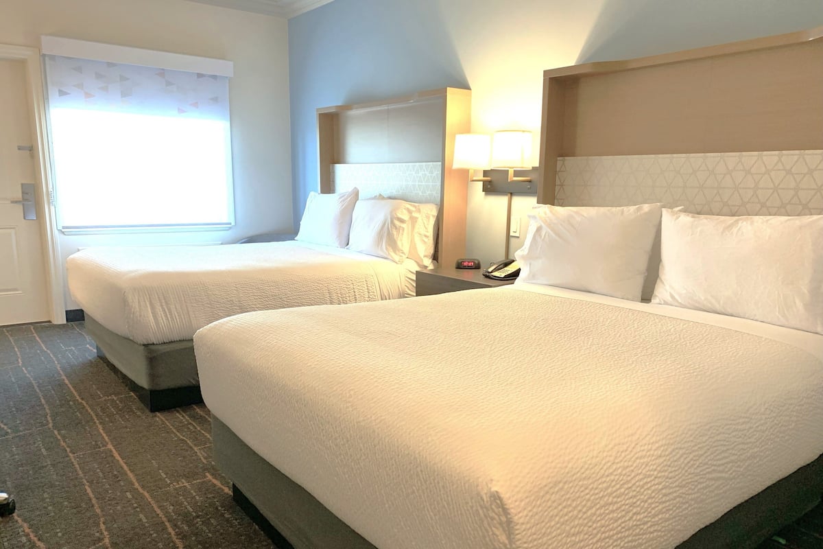 A hotel room with two beds, a nightstand with a lamp, and a window with blinds.