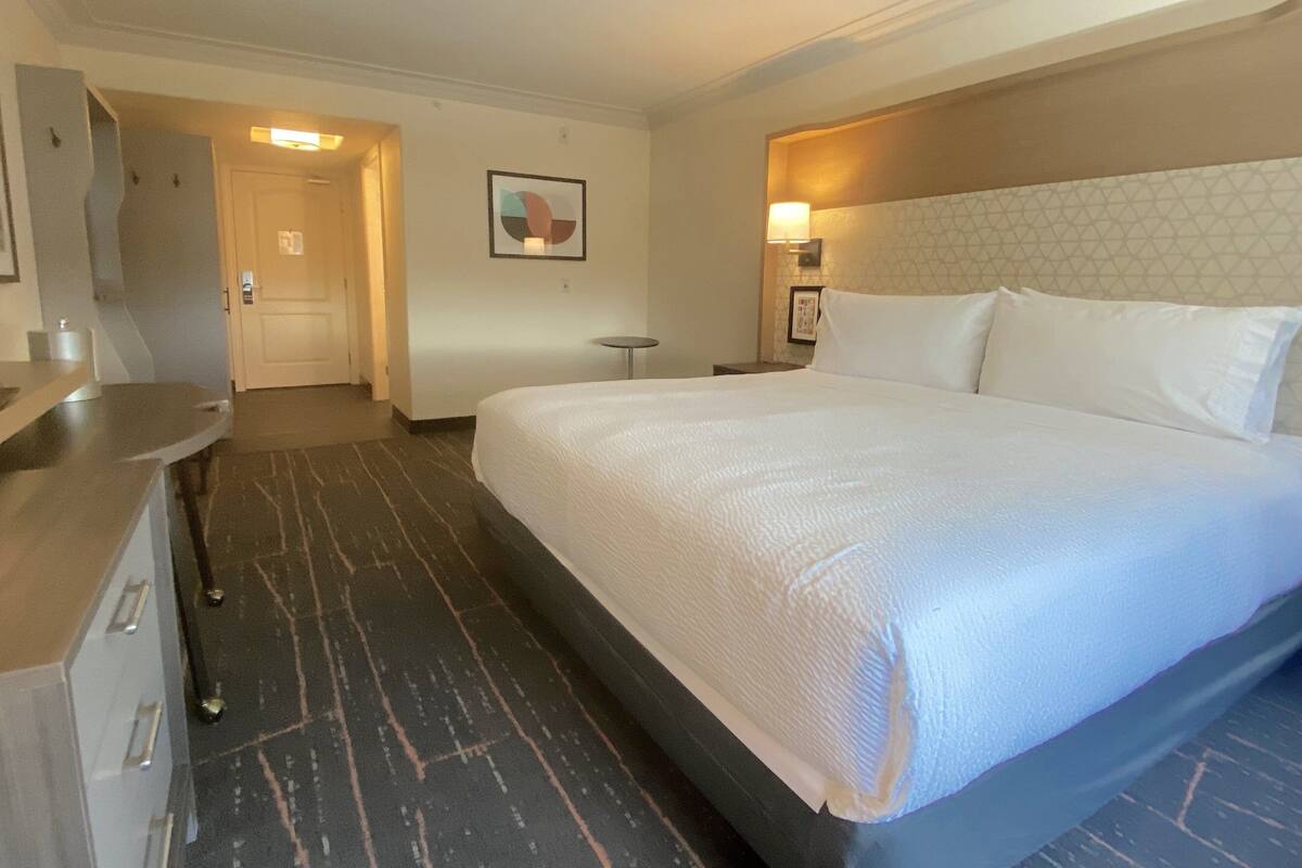 Habitación de hotel con una cama grande, un escritorio, una silla, una mesita y una puerta que da a otra habitación.