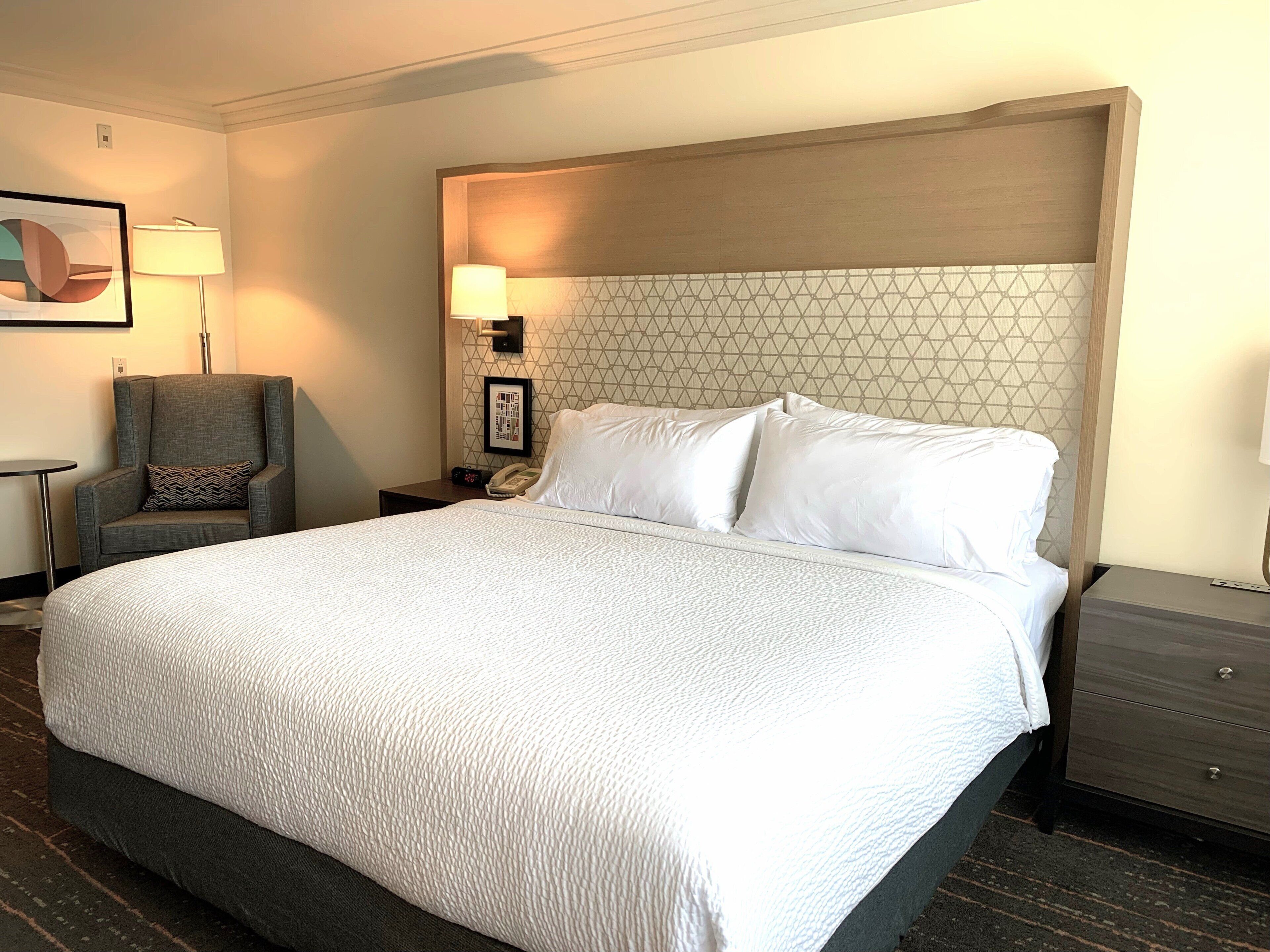 Una habitación de hotel con una cama grande, una silla, una mesita de noche con una lámpara y un cuadro en la pared.